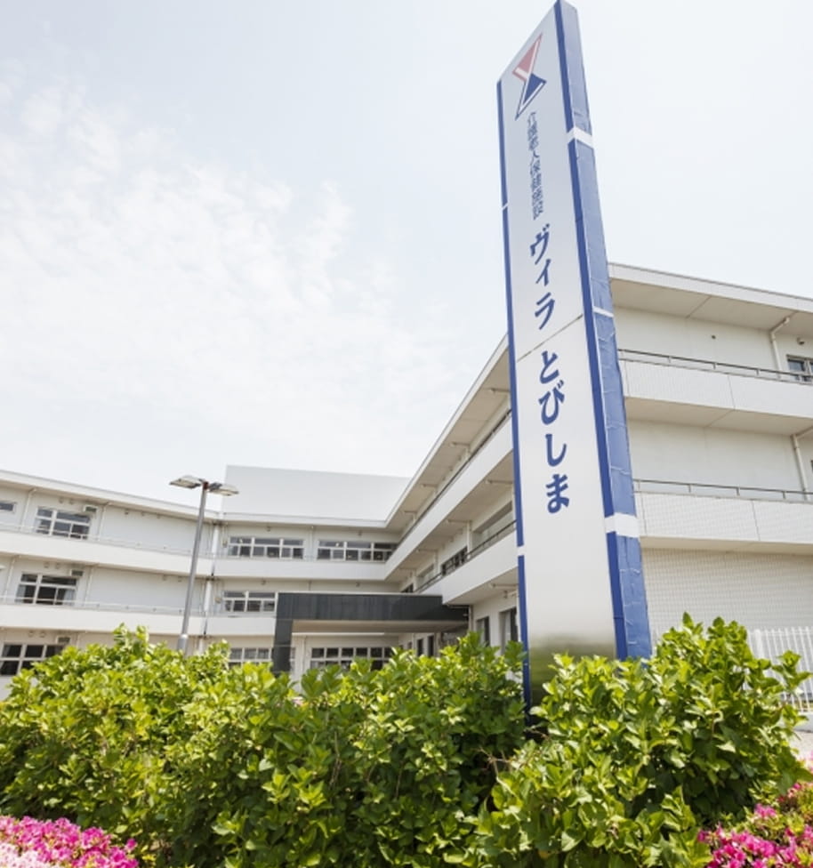 愛知県飛島村の介護老人保健施設ヴィラとびしまの外観。白い多層階の建物と青と白の看板が設置されたモダンな施設。緑豊かな植栽に囲まれた清潔で明るい施設外観