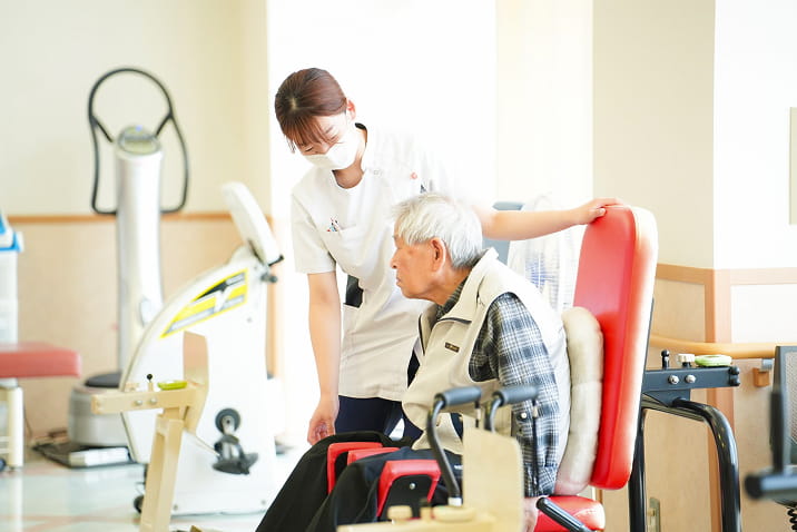 医療法人良斉会ヴィラとびしまのリハビリテーションサポート。スタッフが高齢者の機能訓練を補助し、リハビリテーション機器を使った運動をサポート