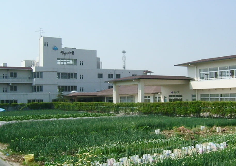 愛知県飛島村の特別養護老人ホームやすらぎの里の外観。手入れされた畑とモダンな白い建物群が広がる開放的な環境。高齢者や地域住民が自然と触れ合える施設外観