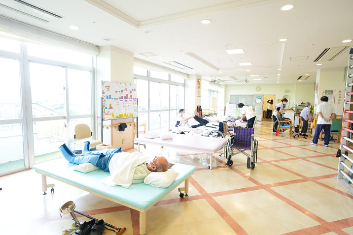 ヴィラとびしまの広々としたリハビリテーション・レクリエーションルーム。自然光が差し込む明るい空間で、複数のベッドや車椅子、リハビリテーション機器が配置された機能訓練室。利用者とスタッフが共に活動するデイサービス施設